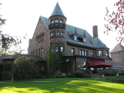 A large, historic stone mansion with a turret, ivy-covered walls, and a green lawn in front.