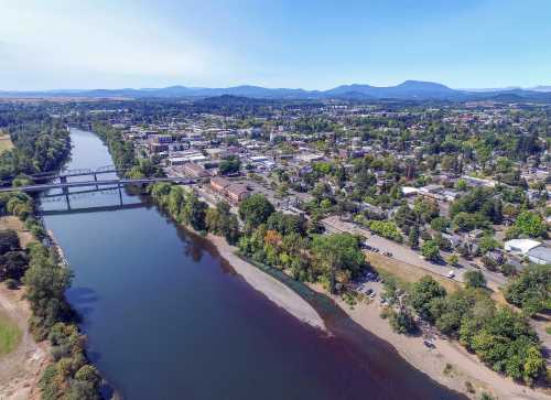 Aerial view of a river winding through a city, surrounded by greenery and mountains in the background.