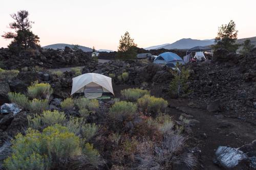 A campsite with tents surrounded by rocky terrain and sparse vegetation, set against a backdrop of mountains at dusk.