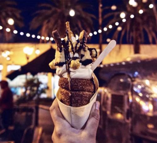 A hand holds a dessert cone filled with soft serve ice cream, topped with chocolate drizzle and crispy treats, under string lights.
