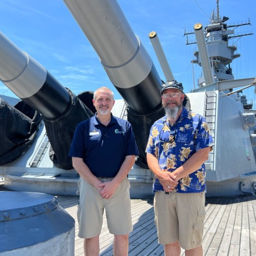 Two men stand on a battleship deck, smiling in front of large naval cannons and ship structures under a clear blue sky.