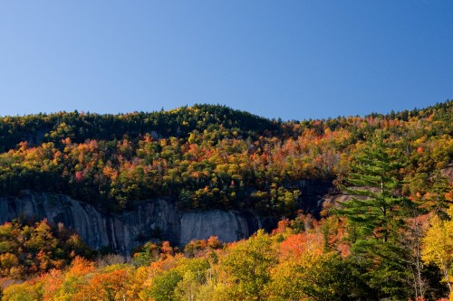 Vibrant autumn foliage covers a mountain, with a clear blue sky above and rocky cliffs in the foreground.
