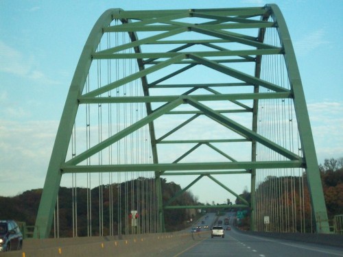 A green arch bridge spans a highway, with cars traveling underneath and trees in the background.