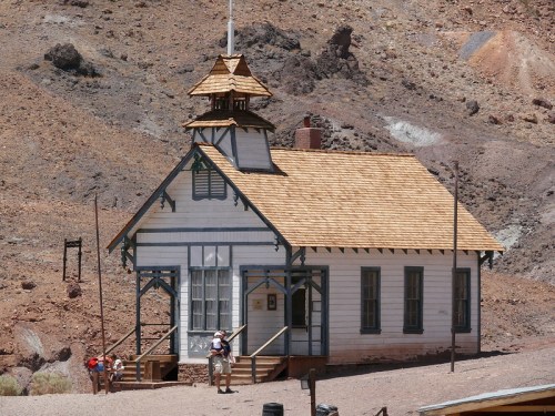 A historic wooden building with a peaked roof, surrounded by rocky terrain and visitors exploring the area.