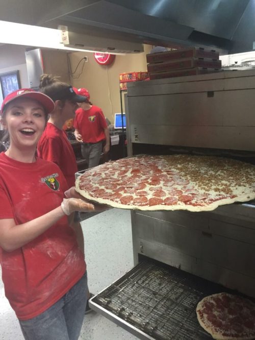A smiling employee holds a large pizza just out of the oven, with others working in the background.