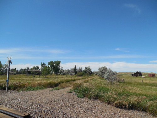 A wide view of a grassy landscape with trees, a dirt path, and distant buildings under a clear blue sky.