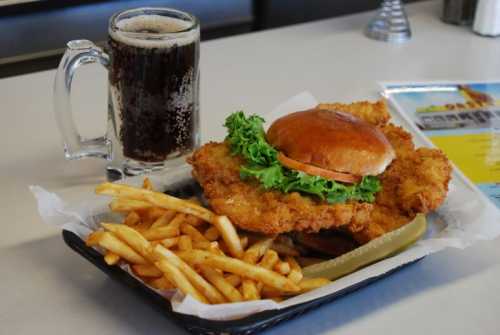 A plate with a large fried chicken sandwich, fries, a pickle, and a glass of soda on a table.