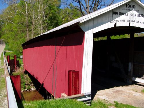 A red covered bridge with a white roof, surrounded by greenery and a gravel path, marked with the year 1900.