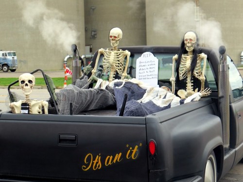 A pickup truck with skeletons in the back, dressed in costumes, surrounded by smoke and a sign that reads "RIP."