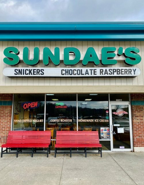 Exterior of a shop named "Sundae's" with a sign featuring flavors like Snickers and Chocolate Raspberry, and red benches outside.