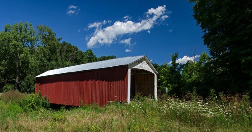 A red barn with a metal roof stands in a field surrounded by greenery and blue skies with fluffy white clouds.