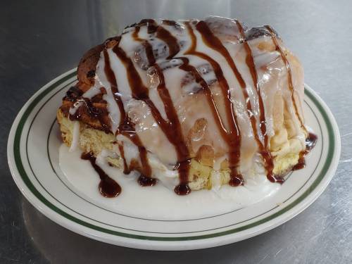 A slice of cinnamon roll topped with icing and chocolate drizzle on a white plate.