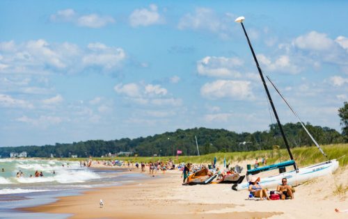 A sunny beach scene with people relaxing, sailboats on the shore, and gentle waves under a blue sky with clouds.