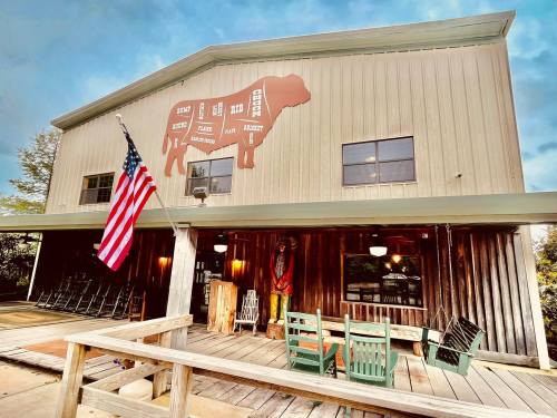 A rustic building with a large cow mural, American flag, and wooden deck with chairs in front.