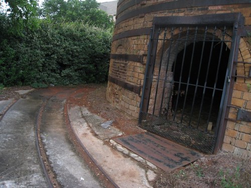 A stone structure with a gated entrance, surrounded by greenery and a curved pathway leading to it.