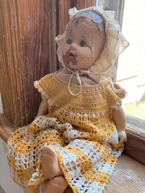 A vintage doll with a cracked face, wearing a yellow and white crocheted dress, sitting by a window.