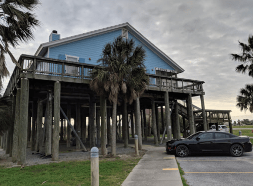 A blue house on stilts with palm trees nearby, parked car in the foreground, under a cloudy sky.