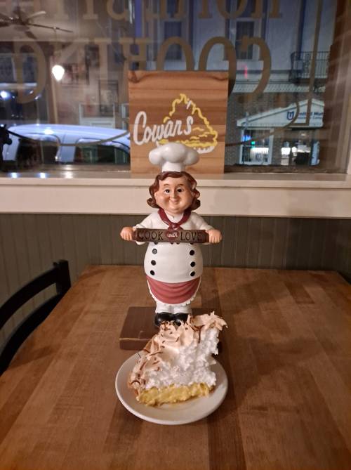 A chef figurine holds a sign reading "Cook with Love" next to a slice of pie topped with meringue on a table.