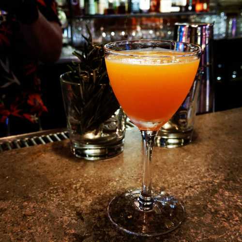 A vibrant orange cocktail in a stemmed glass, sitting on a bar counter with blurred bottles in the background.