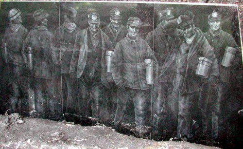 A mural depicting a line of miners in historical clothing, holding lanterns and tools, set against a dark background.
