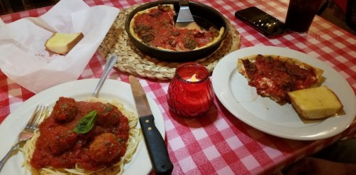 A table with spaghetti and meatballs, deep-dish pizza, garlic bread, and a candle on a red-checkered tablecloth.