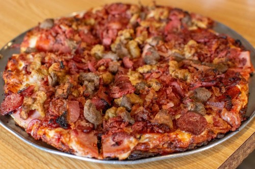 A large pizza topped with various meats, including pepperoni, sausage, and bacon, on a metal serving tray.