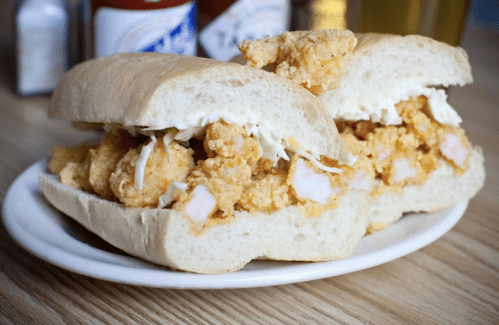 Two fried shrimp sandwiches on a plate, filled with shrimp and topped with coleslaw, with condiments in the background.