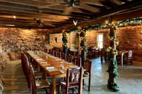 A rustic dining room with a long wooden table, surrounded by stone walls and decorated with string lights and greenery.