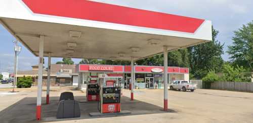 A gas station with fuel pumps and a convenience store, set against a cloudy sky and surrounded by trees.