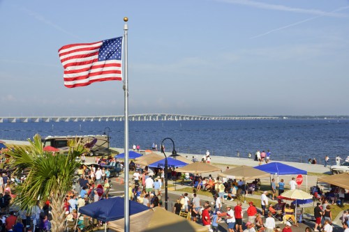 A vibrant waterfront scene with a U.S. flag, crowds, tents, and a bridge in the background under a clear blue sky.