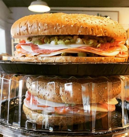A large, stacked sandwich with sesame seed buns, layers of deli meats, pickles, and toppings on a black tray.