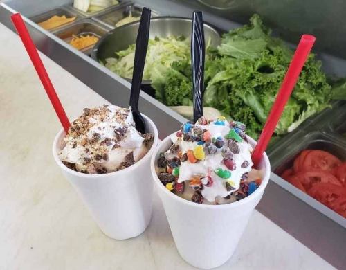 Two cups of ice cream topped with whipped cream, one with chocolate crumbs and the other with colorful candy pieces.