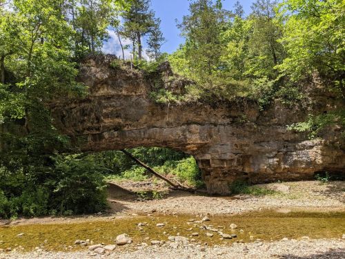 A natural stone arch surrounded by lush greenery and a shallow stream beneath it on a sunny day.