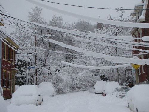 A snowy street scene with heavy snowfall covering cars and power lines, creating a winter wonderland atmosphere.