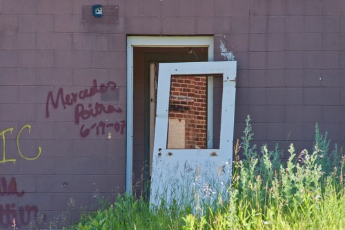 An abandoned building with a partially open door and graffiti on the wall, surrounded by overgrown grass.