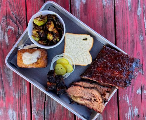 A tray of barbecue featuring brisket, ribs, pickles, onions, a slice of bread, and roasted Brussels sprouts.