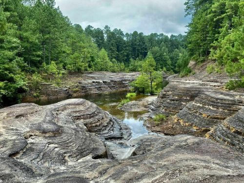A serene landscape featuring layered rock formations and a calm river, surrounded by lush green trees.
