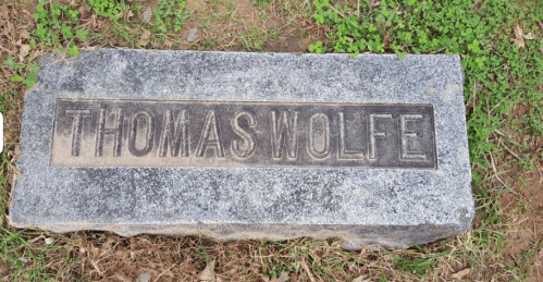 Gravestone with the name "THOMAS WOLFE" engraved on a flat stone, surrounded by grass.