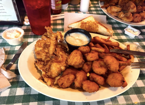 A plate of fried fish, hushpuppies, and French fries, served with a side of dipping sauce and toast on a checkered tablecloth.
