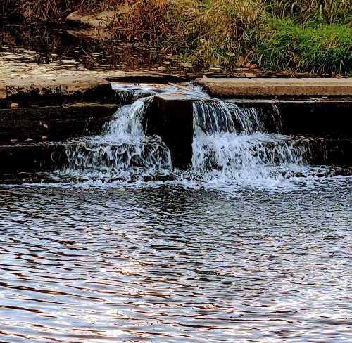 A small waterfall cascades over rocks into a calm body of water, surrounded by grass and natural scenery.