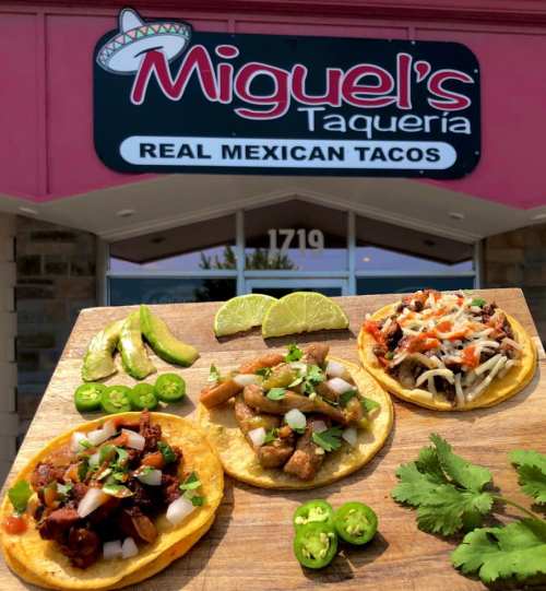 Three tacos on a wooden board, topped with various ingredients, in front of Miguel's Taqueria sign. Fresh limes and jalapeños nearby.