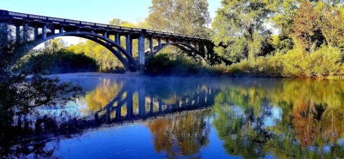A serene river scene with a bridge arching over, surrounded by trees and mist reflecting on the water's surface.