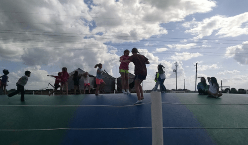 Children playing on a large inflatable bounce pad under a cloudy sky, with some running and others jumping.