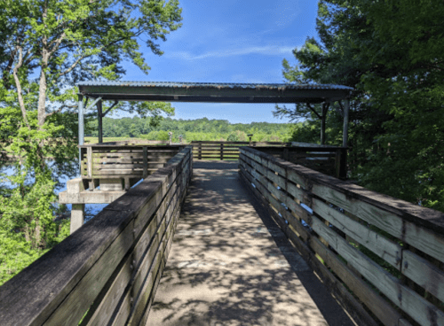 A wooden walkway leads to a covered viewing platform surrounded by trees and a serene lake.