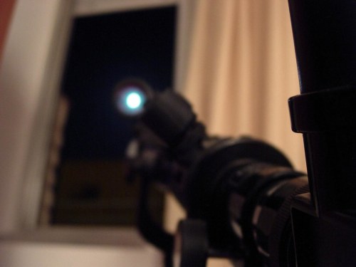 A close-up of a rifle scope aimed out a window, with a bright moon visible in the distance.