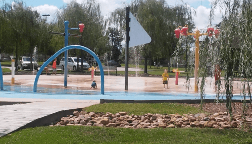 Children playing in a splash pad with colorful water features and a grassy area in a sunny park.