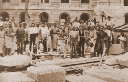 A historic black-and-white photo of a large group of workers posing at a construction site with stone blocks.