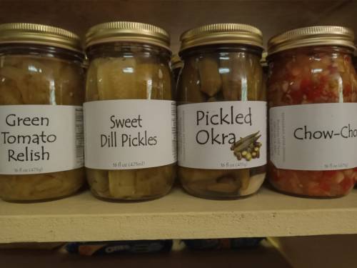 Four jars of pickled products on a shelf: green tomato relish, sweet dill pickles, pickled okra, and chow-chow.