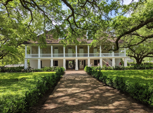 A historic two-story house with a porch, surrounded by lush greenery and trees, leading to a brick pathway.