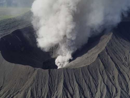 Aerial view of a volcano erupting, with thick smoke and ash billowing from the crater into the sky.